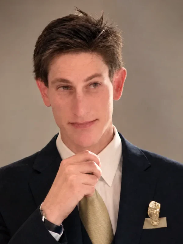 Portrait of author Aaron Holden in a navy suit and gold tie, hand at his chin.